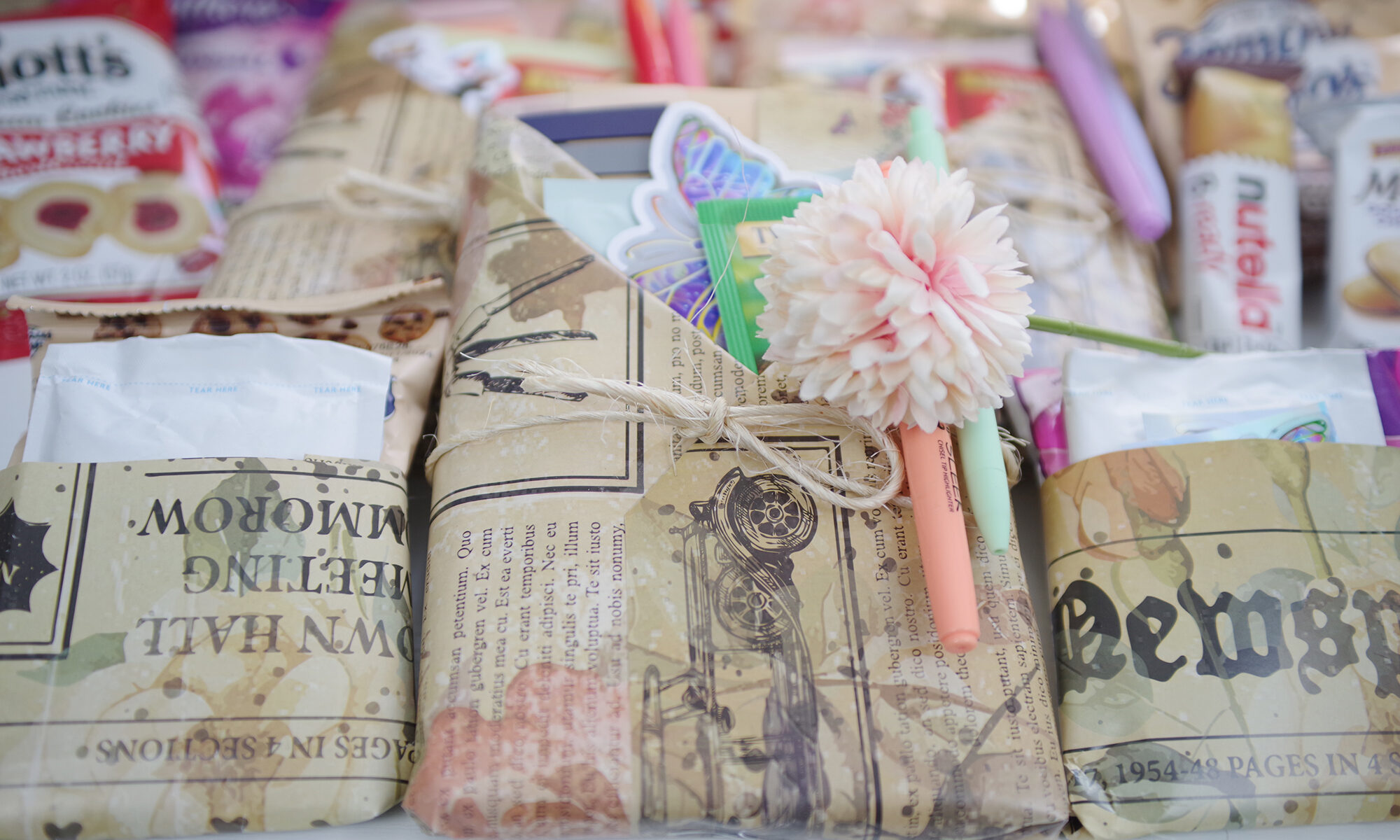 A top down view of a gift wrapped book. The wrapping is done with pockets with the pockets filled with accessories like stickers, tea bags, and pastel highlighters. The image is bright with pastel hues.