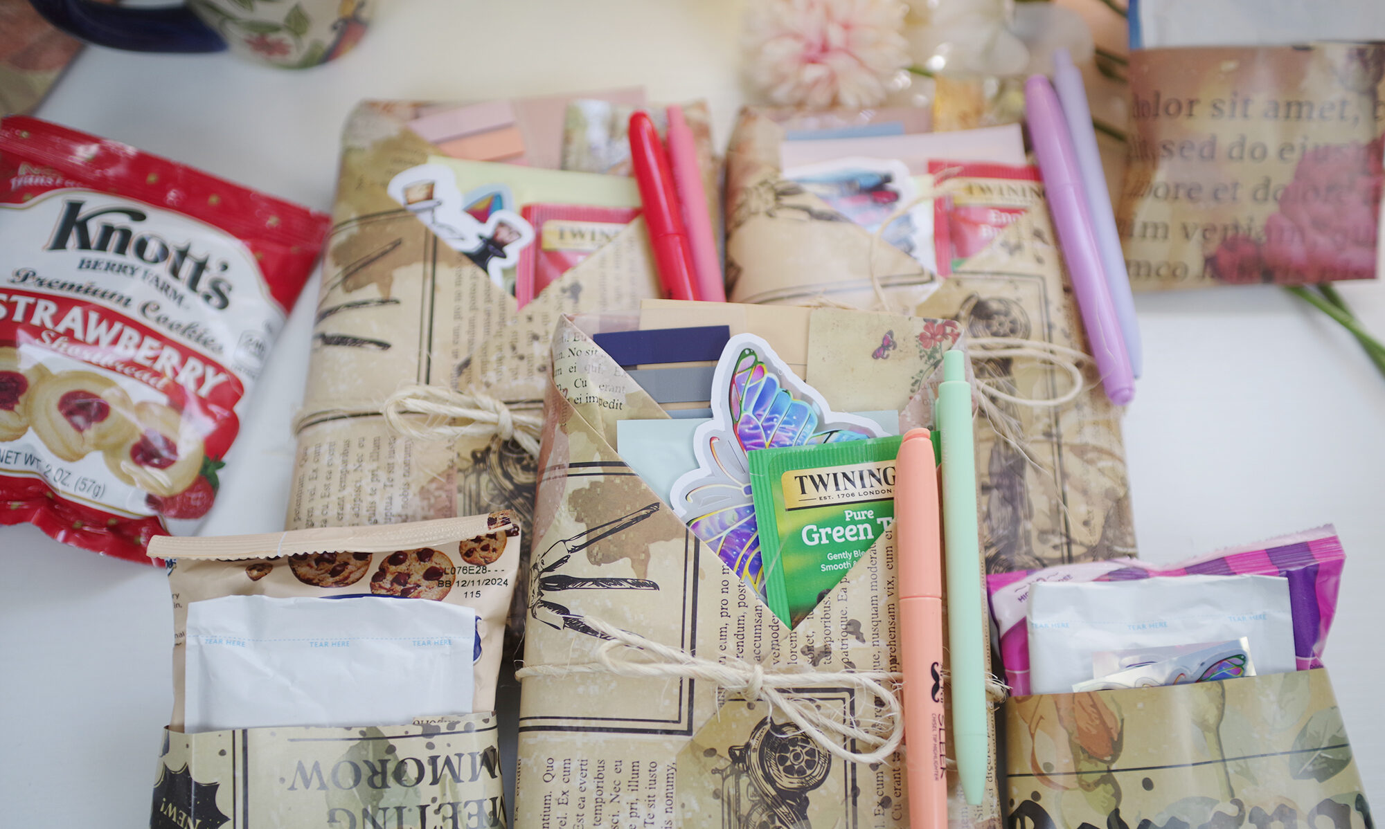 A top down view of a gift wrapped book. The wrapping is done with pockets with the pockets filled with accessories like stickers, tea bags, and pastel highlighters. The image is bright with pastel hues.